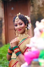 A woman dressed in traditional attire, adorned with intricate gold jewelry and vibrant makeup. Her hair is styled with flowers, and she is wearing a saree with gold embroidery. The background suggests an outdoor setting, possibly near a structure or garden.