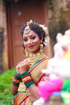 A woman dressed in traditional attire, adorned with intricate gold jewelry and vibrant makeup. Her hair is styled with flowers, and she is wearing a saree with gold embroidery. The background suggests an outdoor setting, possibly near a structure or garden.