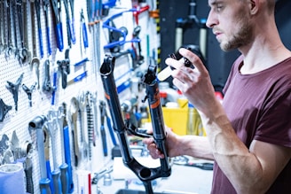 A person is working on a bicycle front suspension fork in a workshop filled with various tools hanging on a wall. The individual appears to be applying lubricant or sealant to the fork using a syringe.