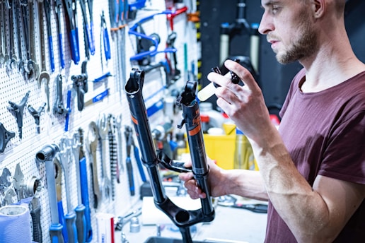 A person is working on a bicycle front suspension fork in a workshop filled with various tools hanging on a wall. The individual appears to be applying lubricant or sealant to the fork using a syringe.
