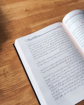 An open book featuring text in Arabic is resting on a wooden surface. Natural light illuminates the page, revealing intricate typographic details.