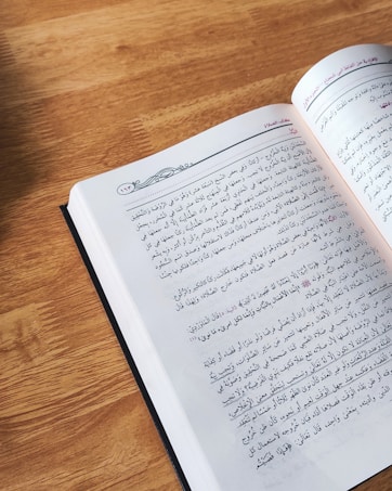 An open book featuring text in Arabic is resting on a wooden surface. Natural light illuminates the page, revealing intricate typographic details.