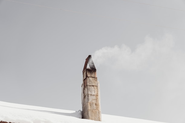 A rustic chimney emits a steady stream of smoke against a pale gray sky, rising from a snow-covered roof.