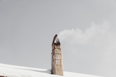 A rustic chimney emits a steady stream of smoke against a pale gray sky, rising from a snow-covered roof.