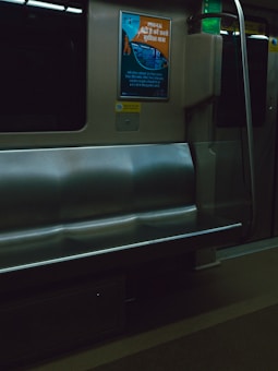 An empty bench seat inside a train or metro car. There is an informational poster on the wall above the seat, written in Hindi, with graphics and text. The environment is metallic, with muted colors and artificial lighting.