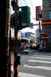 A busy urban street corner with various signage in a mix of Chinese and English. A traffic light pole and pedestrian crossing indicator are in the foreground. In the background, a bus with an advertisement, a yellow taxi, and several people, including a person on a scooter, are visible. The environment suggests an Asian city, with surrounding buildings and overhead cables.
