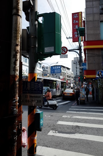 A busy urban street corner with various signage in a mix of Chinese and English. A traffic light pole and pedestrian crossing indicator are in the foreground. In the background, a bus with an advertisement, a yellow taxi, and several people, including a person on a scooter, are visible. The environment suggests an Asian city, with surrounding buildings and overhead cables.