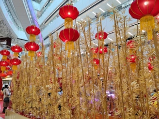 Golden bamboo stalks stretch upwards amidst a modern shopping mall interior, adorned with numerous vibrant red lanterns with yellow tassels. The mall's bright and airy architecture creates a festive atmosphere, enhanced by the traditional decorations.