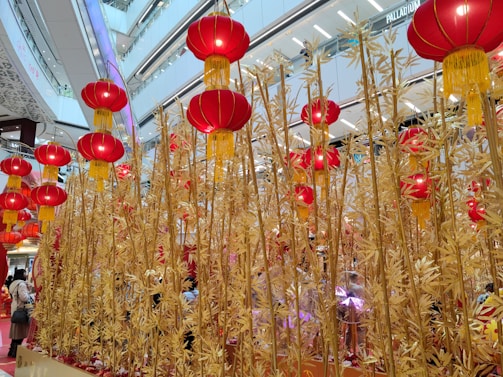 Golden bamboo stalks stretch upwards amidst a modern shopping mall interior, adorned with numerous vibrant red lanterns with yellow tassels. The mall's bright and airy architecture creates a festive atmosphere, enhanced by the traditional decorations.