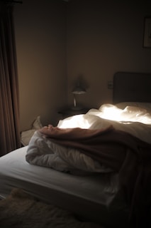 A dimly lit bedroom showcasing an unmade bed with white sheets and a brown blanket. Sunlight streams through the window, casting soft highlights on the bedding. There is a bedside lamp and a small table next to the bed, while the room is adorned with neutral-colored curtains and walls.