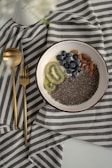 A bowl filled with chia seeds, blueberries, kiwi slices, and almonds is placed on a striped cloth. Beside the bowl, there are gold-colored spoon and fork, adding a touch of elegance. The overall arrangement appears minimalist and aesthetically pleasing.