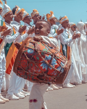 A young child joyfully carries a large, colorful drum while dressed in traditional attire. In the background, a group of people wearing coordinated outfits with orange and green accents clap their hands in unison. The scene suggests a festive or cultural celebration under a clear blue sky.