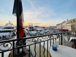 A scenic waterfront view featuring several luxury yachts docked in a marina. The foreground includes a balcony with a round table holding a blue glass and a bottle. People are strolling along the promenade, and there's a line of charming buildings on the right side. The sky is transitioning to sunset, casting a warm glow over the scene.
