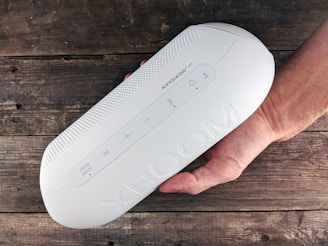 A white, oval-shaped portable speaker labeled 'XBOOM' is held in a person's hand. The speaker features visible control buttons including power, volume, and Bluetooth symbols. The surface has a perforated design on the upper part and is placed against a rustic wooden background.