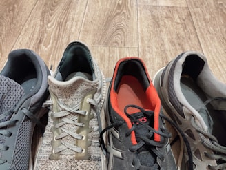 Four different athletic shoes are arranged side by side on a wooden floor. Each shoe is distinct in color and style; the first is a dark gray with geometric mesh patterns, the second has a knit design with shades of beige and green, the third is predominantly black with red accents, and the fourth has a combination of gray and white tones with a striped pattern.