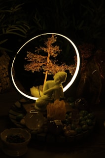 A serene setting featuring a small statue of a kneeling figure in front of a glowing ring with a delicate, illuminated branch inside. The setup is surrounded by various crystals and stones, creating a mystical atmosphere.