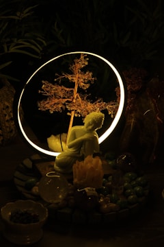 A serene setting featuring a small statue of a kneeling figure in front of a glowing ring with a delicate, illuminated branch inside. The setup is surrounded by various crystals and stones, creating a mystical atmosphere.