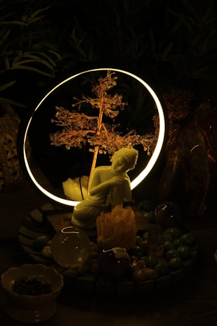 A serene setting featuring a small statue of a kneeling figure in front of a glowing ring with a delicate, illuminated branch inside. The setup is surrounded by various crystals and stones, creating a mystical atmosphere.