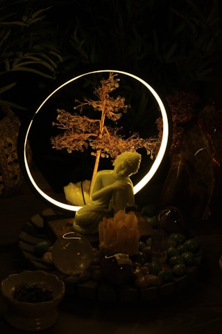 A serene setting featuring a small statue of a kneeling figure in front of a glowing ring with a delicate, illuminated branch inside. The setup is surrounded by various crystals and stones, creating a mystical atmosphere.