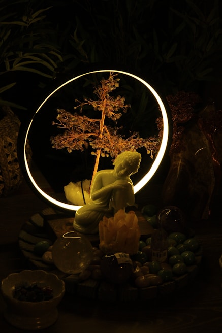 A serene setting featuring a small statue of a kneeling figure in front of a glowing ring with a delicate, illuminated branch inside. The setup is surrounded by various crystals and stones, creating a mystical atmosphere.