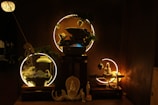 A dimly lit scene showcasing a set of ornamental objects arranged on wooden platforms. In the center, circular light fixtures highlight decor items like a statue of a seated figure and smaller figurines in calming postures. The setup includes elements of greenery and mystical smoke swirling around, creating an ethereal atmosphere.