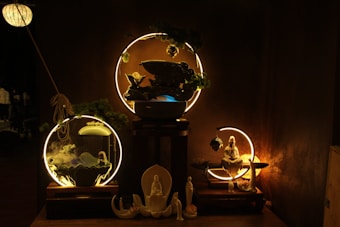 A dimly lit scene showcasing a set of ornamental objects arranged on wooden platforms. In the center, circular light fixtures highlight decor items like a statue of a seated figure and smaller figurines in calming postures. The setup includes elements of greenery and mystical smoke swirling around, creating an ethereal atmosphere.