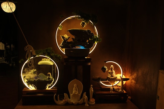 A dimly lit scene showcasing a set of ornamental objects arranged on wooden platforms. In the center, circular light fixtures highlight decor items like a statue of a seated figure and smaller figurines in calming postures. The setup includes elements of greenery and mystical smoke swirling around, creating an ethereal atmosphere.