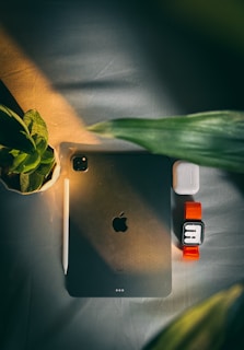 A flat lay composition featuring an Apple iPad with a pencil resting on it, positioned on a light surface. To the right of the iPad, there is a pair of wireless earbuds and a smartwatch with a bright orange strap. Green leaves frame the scene from the left and bottom.