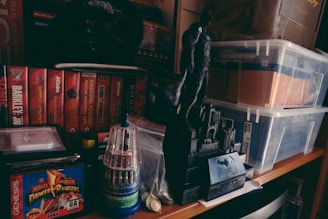 Various Sega Genesis game boxes are arranged neatly on a shelf, including titles like Sonic and Mighty Morphin Power Rangers. A black figurine of a superhero stands prominently on a base designed to look like a cityscape. There are transparent storage boxes filled with files and other items nearby, and a tool set in a clear cylindrical case is also present.