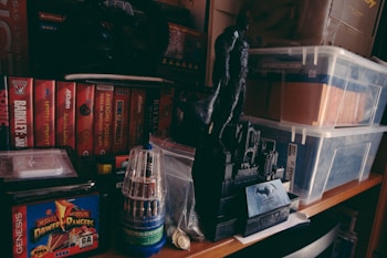 Various Sega Genesis game boxes are arranged neatly on a shelf, including titles like Sonic and Mighty Morphin Power Rangers. A black figurine of a superhero stands prominently on a base designed to look like a cityscape. There are transparent storage boxes filled with files and other items nearby, and a tool set in a clear cylindrical case is also present.