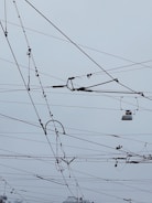 An intricate network of overhead power lines arranged against a cloudy sky. The lines crisscross in various directions, creating a complex pattern. Some parts of the wires seem to include hardware components and insulators.