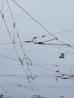 An intricate network of overhead power lines arranged against a cloudy sky. The lines crisscross in various directions, creating a complex pattern. Some parts of the wires seem to include hardware components and insulators.