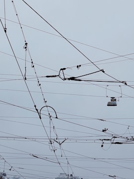 An intricate network of overhead power lines arranged against a cloudy sky. The lines crisscross in various directions, creating a complex pattern. Some parts of the wires seem to include hardware components and insulators.