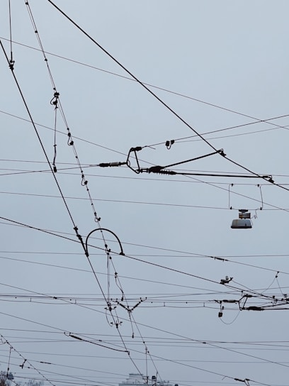 An intricate network of overhead power lines arranged against a cloudy sky. The lines crisscross in various directions, creating a complex pattern. Some parts of the wires seem to include hardware components and insulators.