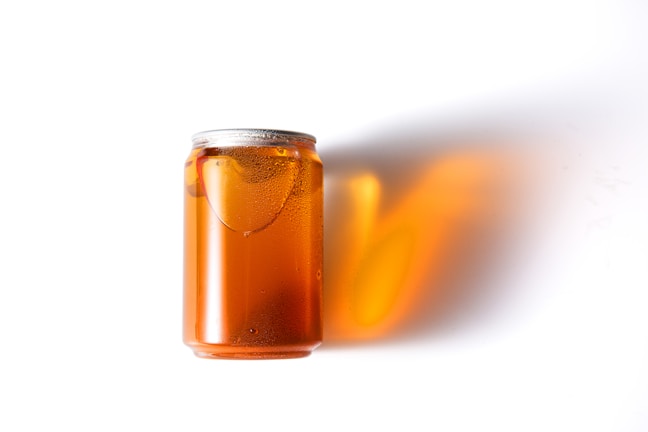 A can filled with amber liquid, likely a beverage, is placed on a white background. The can is partially covered with condensation, indicating that it is cold. The liquid casts a warm, orange-hued shadow on the surface beside the can.