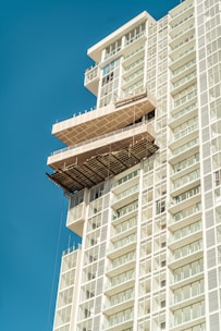 A modern high-rise building featuring a prominent structure protruding from its facade, resembling a large balcony or terrace. The building is composed of multiple rectangular glass windows, and the sky is clear and blue, enhancing the architectural design.