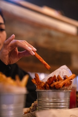 A hand reaching for a sweet potato fry from a small metal bucket filled with fries. The background is blurred, suggesting a cozy indoor setting with warm lighting.