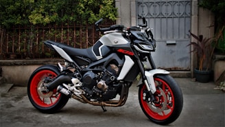 A modern, sporty motorcycle with a sleek design, featuring a black and white color scheme with vibrant red wheels, parked in a driveway. The background includes a metal gate and some plants, contributing to an urban setting.