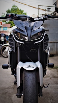A motorcycle with a bold and aggressive design is parked in an outdoor setting. The front view showcases distinctive angular headlights and a prominent wheel with a visible tread. The Yamaha branding is clearly displayed on the front fender. The background features a blurred mix of greenery and urban elements.