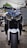 A motorcycle with a bold and aggressive design is parked in an outdoor setting. The front view showcases distinctive angular headlights and a prominent wheel with a visible tread. The Yamaha branding is clearly displayed on the front fender. The background features a blurred mix of greenery and urban elements.
