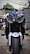 A motorcycle with a bold and aggressive design is parked in an outdoor setting. The front view showcases distinctive angular headlights and a prominent wheel with a visible tread. The Yamaha branding is clearly displayed on the front fender. The background features a blurred mix of greenery and urban elements.