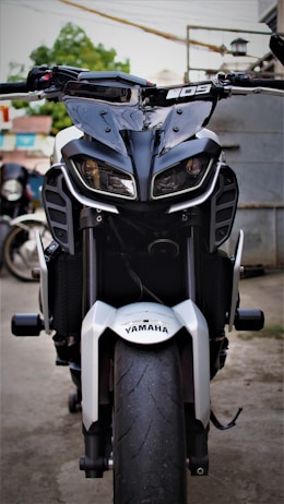 A motorcycle with a bold and aggressive design is parked in an outdoor setting. The front view showcases distinctive angular headlights and a prominent wheel with a visible tread. The Yamaha branding is clearly displayed on the front fender. The background features a blurred mix of greenery and urban elements.