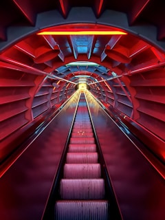 A futuristic escalator is enclosed within a tunnel-like structure, illuminated by vibrant red and blue lights. The tunnel's walls have a repetitive, symmetrical pattern, enhancing the feeling of depth and movement as the escalator ascends towards a bright, glowing exit.