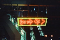 A neon sign with red characters framed in yellow hangs in an industrial or urban setting. The background is dark, with glimpses of metal structures and faint light sources. The scene evokes a night-time or indoor environment with an ambient glow.
