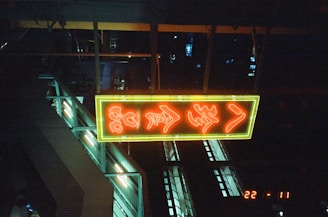 A neon sign with red characters framed in yellow hangs in an industrial or urban setting. The background is dark, with glimpses of metal structures and faint light sources. The scene evokes a night-time or indoor environment with an ambient glow.