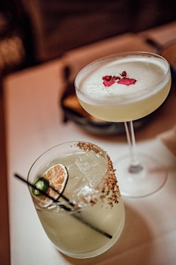 Two elegant cocktails presented on a table. One is a margarita glass filled with a frothy, light-yellow liquid garnished with red flower petals. The other is a short glass with a salted rim, containing ice, a slice of lime, and a green chili pepper, likely a margarita.