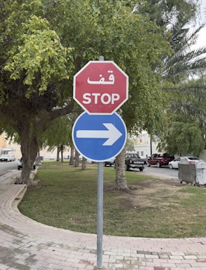 A red octagonal stop sign with Arabic and English text is combined with a blue circular sign indicating a right turn. The signs are placed on a grassy area next to a sidewalk, surrounded by trees. Cars are parked along the street in the background.