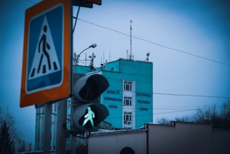 A teal-colored building with signage in a foreign language is in the background, with antennas and satellite dishes on the roof. In the foreground, there is a pedestrian traffic light displaying a green signal, and a pedestrian crossing sign mounted on a pole.