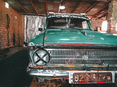 An old teal-colored car with visible wear and rust sits inside a rustic garage. Its front grill features a round headlight with a black tape 'X' over it on one side. The brick walls and wooden ceiling structure of the garage provide a weathered backdrop, and there's a corrugated metal sheet partially covering an entrance or window.