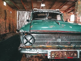 An old teal-colored car with visible wear and rust sits inside a rustic garage. Its front grill features a round headlight with a black tape 'X' over it on one side. The brick walls and wooden ceiling structure of the garage provide a weathered backdrop, and there's a corrugated metal sheet partially covering an entrance or window.